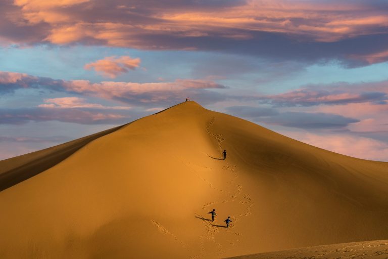 Three people ascend a sunlit sand dune under a vibrant sky, creating a serene desert scene.